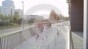 Blonde woman and shirtless man jog up a sloping pavement towards green avenue.