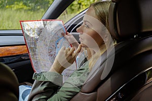 Blonde woman sitting inside the car checking the route on map to get to the destination. Afraid to get lost in forest