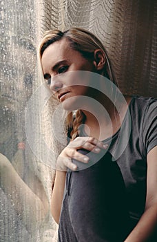 Blonde woman sitting by the rainy window, looking absently out, sad