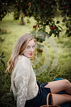 Blonde woman is sitting under a tree full of red apples. Wearing a cozy sweater and skirt, enjoying warm autumn evening in garden