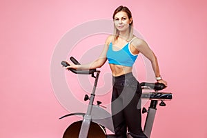 Blonde woman with slim body shape posing near exercise bike and looking at camera.
