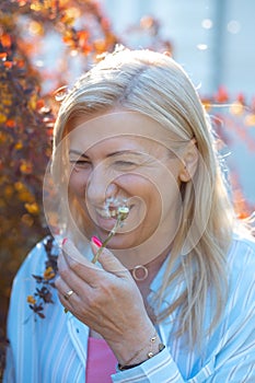 Blonde woman smiling shyly. In hand of lady white dandelion which has just blow off. Happy time outdoor in summer.