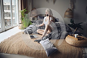 Blonde woman speaking phone working, making video call, watching movie on a laptop in cozy home bedroom modern interior.