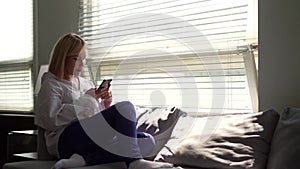 Blonde woman is spending time in social networks, using smartphone, staying at home