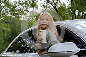 Blonde woman sticking head out of windshield car and drink coffee or tea from reusable mug. Young tourist explore local