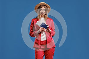 Blonde woman in straw hat, white blouse and red pantsuit. She smiling, holding passport and ticket while posing on blue