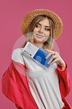 Blonde woman in straw hat, white blouse and red pantsuit. She smiling, holding passport and ticket while posing on pink