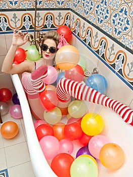 Blonde woman with sunglasses playing in her bath tube with bright colored balloons. Sensual girl with white red striped stockings