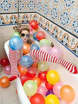 Blonde woman with sunglasses playing in her bath tube with bright colored balloons. Sensual girl with white red striped stockings