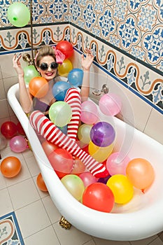 Blonde woman with sunglasses playing in her bath tube with bright colored balloons. Sensual girl with white red striped stockings