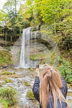 Blonde woman taking a picture of the Cascade du Dard Nozon Gorge, near Croy-RomainmÃ´tier, Canton Vaud, Switzerland