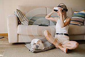 Blonde woman testing VR glasses headset with sleeping small dog.