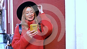 Blonde woman typing and reading text messages on her smartphone in front of a red wall.