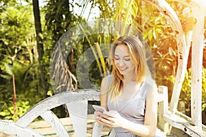 Blonde woman typing smartphone for work on sunny day, background of sunshine green palms in Thailand, Phuket travel