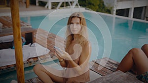 A blonde woman uses her mobile phone while lounging in the sun by the pool on vacation