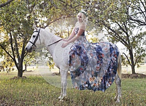 Blonde woman wearing blue floral dress seated on white horse in woods