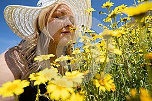 Blonde woman wearing straw white hat smelling a field of yellow wildflowers. Concept for springtime allergy relief