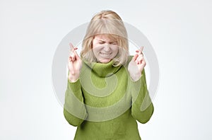 Blonde woman wears green sweater, smiles broadly, keeps fingers crossed, hopes for good luck.