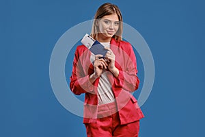 Blonde woman in white blouse and red pantsuit. She smiling, holding passport and ticket while posing on blue studio