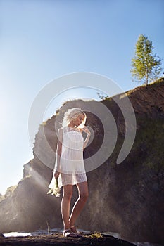 Blonde woman in a white dress and flowers daisies in her hand standing on the rocks near the waterfall. A girl in a light white