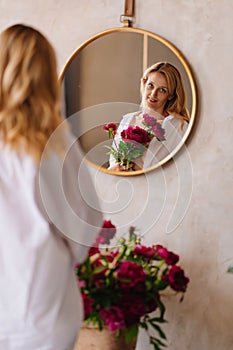 a blonde woman in a white shirt and a bouquet of peonies looks at in the mirror.