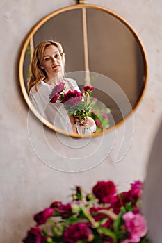 a blonde woman in a white shirt and a bouquet of peonies looks at in the mirror.