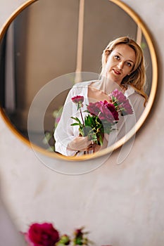 a blonde woman in a white shirt and a bouquet of peonies looks at in the mirror.