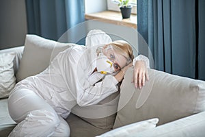 Blonde woman in white workwear and respirator resting on the sofa