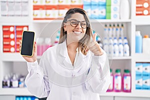 Blonde woman working at pharmacy drugstore showing smartphone screen smiling happy and positive, thumb up doing excellent and