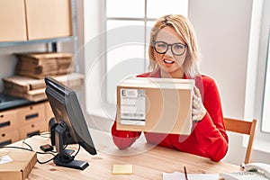 Blonde woman working at small business ecommerce holding box clueless and confused expression