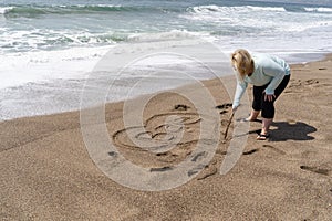 Blonde woman writes Bae slang for Before Anyone Else, representing a romantic relationship in sand on the beach