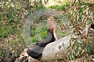 Blonde, young and beautiful woman dressed in black and sitting on the trunk of a fallen eucalyptus tree in the forest, the woman
