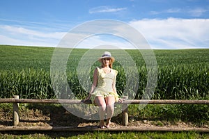Blonde, young, pretty farm woman, sitting on a wooden fence. In the background a green wheat field. The woman is wearing a short