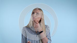 Blonde young woman holding her chin with hand while thinking about something on blue background.