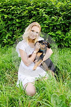 A  blonde young woman sitting in green grass holds dog in her arms.Vertical view