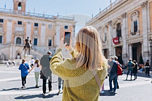 Blonde young Woman with smartphone is walking on a sunny day. Capitol in Rome, Piazza del Campidoglio in Capitoline Hill