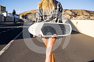 Blonde young woman viewed from rear walking down the street in a little city near a mountain with a skateboard. youth and