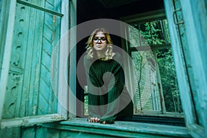 A blonde young woman in a vintage green sweater stands looking out of a broken window in an old abandoned house. Loneliness