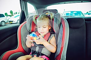 Blondy baby girl fastened with security belt in safety car seat and plaing with toy. Child in auto baby seat in car. Transport, sa