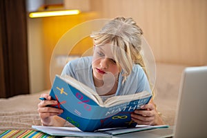 A blone girl lying on the bed and doing her lessons