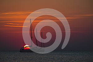 Bloody red sun setting down behind the silohouettes of a two sailboats or yachts moored on an open sea. Adventourous sunset