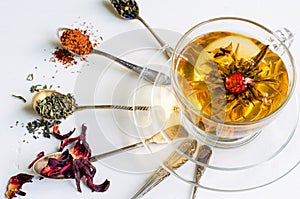 Blooming or flowering tea in a glass cup and spoons with various kinds of tea on white background