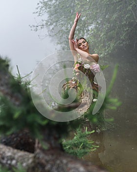 Blooming gorgeous lady in dress of flowers by the lake in the forest in the morning fog