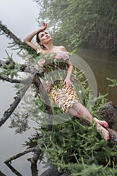 Blooming gorgeous lady in dress of flowers lying on tree over water in the morning fog