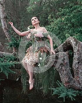 Blooming gorgeous lady in dress of flowers sitting on tree over water in the morning fog