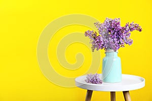 Blossoming lilac flowers in vase on table against color background.