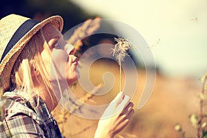 Blowing away her cares.... Beautiful young woman wearing a straw fedora and blowing at a dandelion.