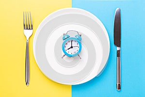 Blue alarm clock, fork, knife and empty plate on colored paper background. Intermittent fasting concept