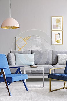 Blue armchair near white table and grey sofa in modern living room interior with poster. Real photo