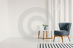 Blue armchair next to white round coffee table with vase with flowers in empty white interior with striped rug, real